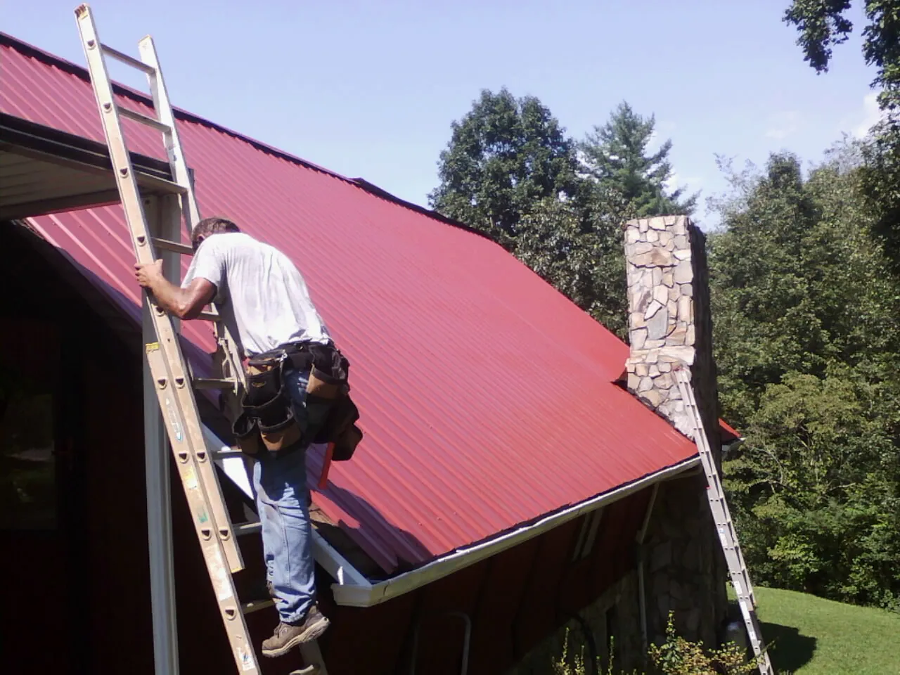 Roofer on a ladder beside a red metal roof and stone chimney — Soffit & Fascia Repair by Roofing Brothers in Scotts